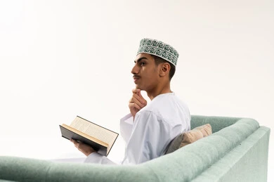 ممارسة هواية المطالعة, مفهوم الثقافة والتعلم الذاتي, الاستمتاع بالأنشطة الترفيهية المفيدة في وقت الفراغ, قراءة القصص والروايات, بورتريه من الخلف لشاب عربي خليجي عماني يرتدي الدشداشه و الكمه يجلس على الاريكة يحمل بيده كتاب و ينظر الى شيء ما بايماءات التفكير, خلفية بيضاء