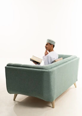 مفهوم الثقافة والتعلم الذاتي، ممارسة هواية المطالعة, الاستمتاع بالأنشطة الترفيهية المفيدة في وقت الفراغ, قراءة القصص والروايات, بورتريه من الخلف لشاب عربي خليجي عماني يرتدي الدشداشه و الكمه يجلس على الاريكة يحمل بيده كتاب و ينظر الى شيء ما بايماءات التفكير, خلفية بيضاء