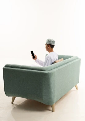 Chatting and talking with friends online, browsing mobile applications, a portrait from behind of a young Arab Gulf Omani man wearing a dishdasha and kumma sitting on the sofa following social media news using his mobile phone, white background.