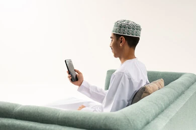 تصفح تطبيقات الهاتف المحمول، الدردشة والتحدث مع الاصدقاء عبر الانترنت، بورتريه من الخلف لشاب عربي خليجي عماني يرتدي الدشداشه و الكمه يجلس على الاريكة يقوم بمتابعة اخبار وسائل التواصل الاجتماعي باستخدام الجوال، خلفية بيضاء