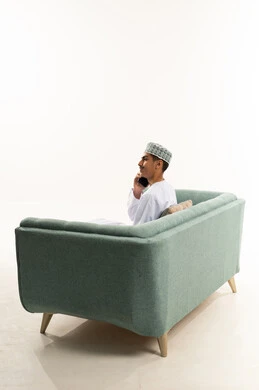 Communication with family and friends, a portrait from behind of an Arab Gulf Omani man wearing a dishdasha and kumma sitting on the sofa making a phone call via a mobile phone, using advanced modern technologies, white background.