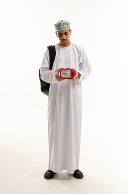 The importance of waking up early to go to school, a portrait of an Arab Gulf Omani student wearing a dishdasha and kumma, carrying a school backpack, holding an alarm clock in his hand and looking at it, back-to-school season in the Sultanate of Oman, full-body shot, white background.