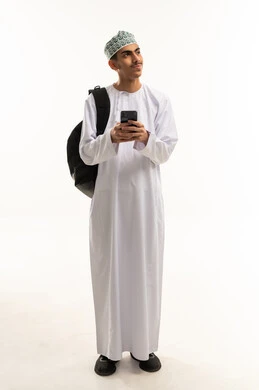 Looking at something, chatting with friends online, a portrait of an Arab Gulf Omani student wearing a dishdasha and kumma carrying a bag on his back, following social media news using a mobile phone, browsing mobile applications, full-body image, white background. Looking at something, chatting with friends online, a portrait of an Arab Gulf Omani student wearing a dishdasha and kumma carrying a bag on his back, following social media news using a mobile phone, browsing mobile applications, full-body image, white background.