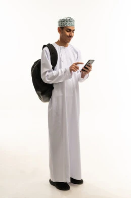الدردشة مع الاصدقاء عبر الانترنت، بورتريه لطالب عربي خليجي عماني يرتدي الدشداشة و الكمة يحمل على ظهره حقيبه يتابع اخبار وسائل التواصل الاجتماعي باستخدام الجوال، تصفح تطبيقات الهاتف المحمول، ايماءات الاعجاب والسرور، صورة الجسم بالطول الكامل، خلفية بيضاء