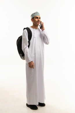 دمج التقنية والتكنولوجيا في الحياة اليومية والعملية، بورتريه لطالب ثانوي عربي خليجي عماني يرتدي الدشداشة و الكمة و حقيبة الظهر يقوم بأجراء مكالمة هاتفية باستخدام الهاتف المحمول، التواصل مع الاهل والاصدقاء، الجسم بالطول الكامل، خلفية بيضاء