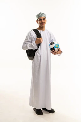 Looking at the camera with gestures of happiness and joy, cheering for the football team, a portrait of an Arab Gulf Omani student wearing a dishdasha and kumma, carrying a bag on his back and holding a football in his hand, full-body, white background.