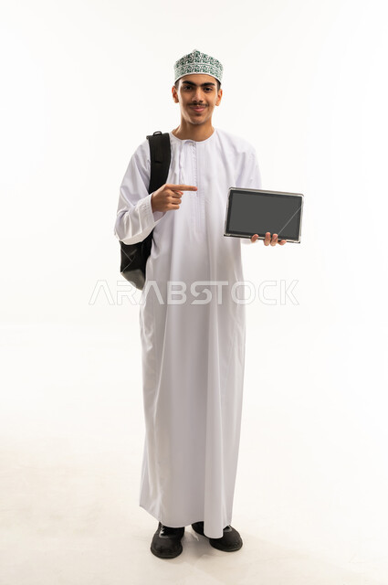 The concept of distance education, educational materials and curricula in the Sultanate of Oman, following lectures through advanced educational platforms, a portrait of an Omani Gulf Arab high school student wearing a dishdasha and kumma, carrying a bag on his back, holding a tablet in his hands and pointing to it, full-body image, white background.