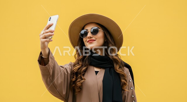 استخدام الهاتف المحمول بايماءات السعادة والفرح، الاهتمام بالاناقة والمظهر الخارجي، بورتريه مقرب لإمرأة عربية خليجية سعودية ترتدي الزي الكاجوال وقبعة القش تضع النظارات الشمسية تقوم بالتقاط صور سيلفي شخصية، خلفية صفراء