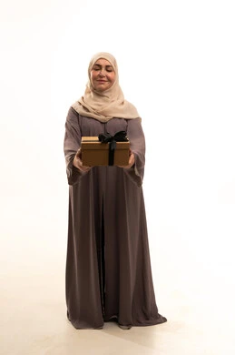 تقديم الهدايا في المناسبات السعيدة، التعبير عن المحبة والتقدير، الاحتفال بأعياد الميلاد, بورترية لإمراة كبيرة بالسن عربية سعودية خليجية محجبة ترتدي العباءة تمسك بيديها علبة هداية, الجسم بالطول الكامل، خلفية بيضاء