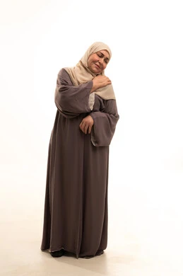 The feeling of illness and fatigue, inflammation of the muscles and joints, the need for treatment and seeking help, a portrait of an elderly Arab Saudi Gulf woman wearing a hijab and an abaya, placing her hand on her shoulder with gestures indicating severe pain, full-body image, white background.