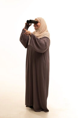 رؤية الأشياء البعيدة، حب الاستكشاف والمغامرة، بورترية لإمراة كبيرة بالسن عربية سعودية خليجية محجبة ترتدي العباءة تمسك منظار ثنائي العينين تشير بإصبع السبابة إلى شيء ما، التخطيط المستقبلي للأهداف والطموحات، الجسم بالطول الكامل، خلفية بيضاء