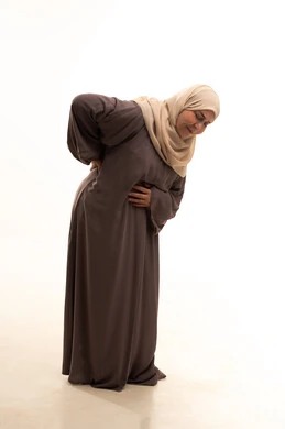 الشعور بالمرض والإرهاق، تعابير الألم ووجع الظهر، بورترية لإمراة كبيرة بالسن عربية سعودية خليجية محجبة ترتدي العباءة تضع يدها على ظهرها بايماءات التعب، الحاجة للعلاج والاستشارة الطبية، الجسم بالطول الكامل، خلفية بيضاء