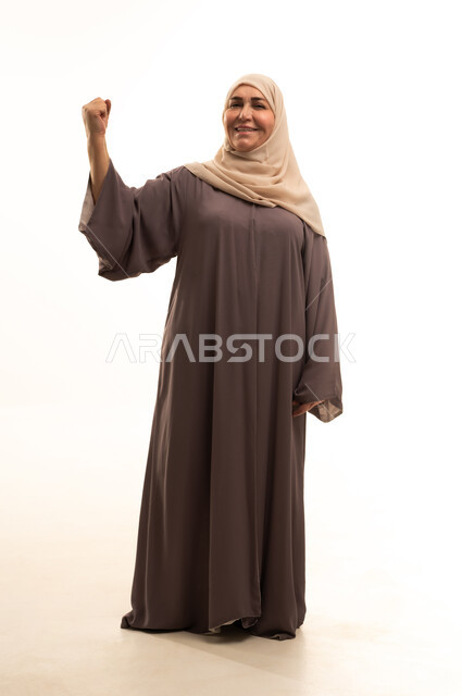 الفرح بالنصر والفوز، تحقيق الأهداف والنجاح، بورترية لإمراة كبيرة بالسن عربية سعودية خليجية محجبة ترتدي العباءة ترفع قبضة يديها بإيماءات الحماس والانتصار، تعابير السعادة و السرور، الجسم بالطول الكامل، خلفية بيضاء