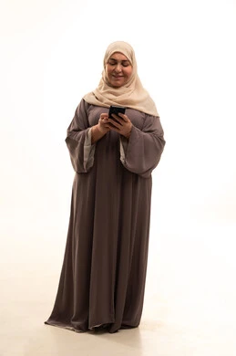 استخدام جهاز تقني حديث ومتطور، تصفح مواقع التواصل الاجتماعي، الدردشة مع الأصدقاء والأقارب عبر الجوال، بورترية لإمراة كبيرة بالسن عربية سعودية خليجية محجبة ترتدي العباءة تمسك بيدها الهاتف المحمول، الجسم بالطول الكامل، خلفية بيضاء