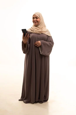 Browsing social media, using a modern and advanced technical device, chatting with friends and relatives via mobile, a portrait of an elderly Arab Saudi Gulf woman wearing a hijab and an abaya holding a mobile phone in her hand, full-body, white background.