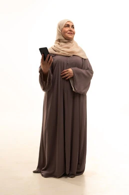 Looking at something, browsing social media, using a modern and advanced technical device, chatting with friends and relatives via mobile, a portrait of an elderly Arab Saudi Gulf woman wearing a hijab and an abaya holding a mobile phone in her hand, full body, white background.