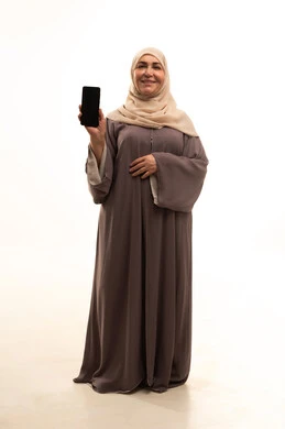Integrating technology and tech into practical life, displaying a blank black screen on a mobile phone, accomplishing tasks quickly and easily, a portrait of an elderly Arab Saudi Gulf woman wearing a hijab and an abaya, holding a mobile phone in her hand, looking at the camera with gestures of happiness and joy, full-body shot, white background.