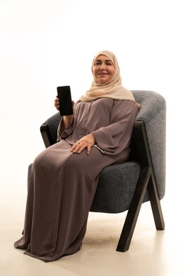 Use modern and advanced devices and technologies, a portrait of an elderly Arab Saudi Gulf woman wearing a hijab and an abaya sitting on a chair displaying a blank black screen on her mobile phone, looking at the camera with gestures of happiness and joy, white background.