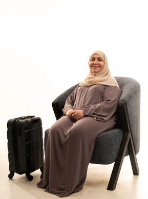 تعابير وايماءات الفرح و السرور، الاستعداد لقضاء رحلة صيفية ممتعة, بورترية لإمراة كبيرة بالسن عربية سعودية خليجية محجبة ترتدي العباءة تجلس على كرسي بجانب حقيبة سفر , خلفية بيضاء