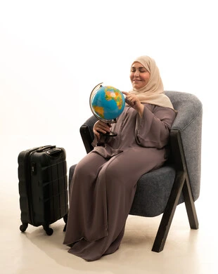 تعابير السرور و الفرح بالانتماء للدول العربيه، مفهوم حب البحث واستكشاف القارات في الجغرافيا, بورترية لإمراة كبيرة بالسن عربية سعودية خليجية محجبة ترتدي العباءة تجلس على كرسي بجانب حقيبة سفر تحمل مجسم الكرة الارضية, خلفيه بيضاء
