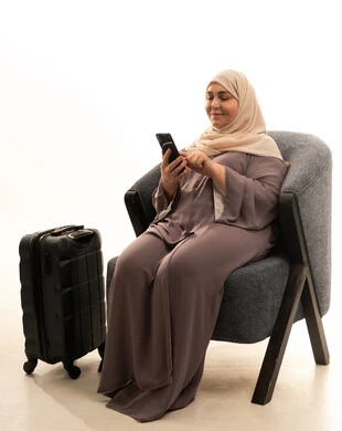 Using modern technologies, communicating with friends via video phone calls, a portrait of an elderly Arab Saudi Gulf woman wearing a hijab and an abaya sitting on a chair next to a suitcase holding a mobile phone in her hands, planning to enjoy the holiday, making travel reservations, white background.