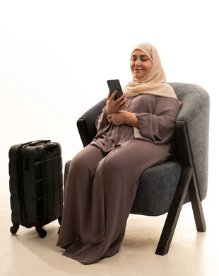 التواصل مع الأصدقاء عبر مكالمة الفيديو الهاتفية ,استخدام التقنيات الحديثة , بورترية لإمراة كبيرة بالسن عربية سعودية خليجية محجبة ترتدي العباءة تجلس على كرسي بجانب حقيبة سفرتمسك بيديها الجوال , التخطيط للاستمتاع بالعطلة , القيام بحجز رحلات السفر , خلفية بيضاء