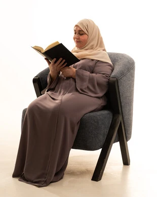 قضاء الوقت بالتعلم والتثقف، ايماءات التأمل والتمعن، ثقافة التعلم الذاتي، الاستمتاع بقراءة الكتب والقصص والروايات، بورترية لإمراة كبيرة بالسن عربية سعودية خليجية محجبة ترتدي العباءة تجلس على كرسي تحمل بيديها كتاب، خلفية بيضاء