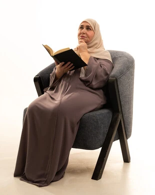 النظر الى مكان ما بإيماءات تذكير شيء ما، ثقافة التعلم الذاتي، قضاء الوقت بالتعلم والتثقف، الاستمتاع بقراءة الكتب والقصص والروايات، بورترية لإمراة كبيرة بالسن عربية سعودية خليجية محجبة ترتدي العباءة تجلس على كرسي تحمل بيديها كتاب، خلفية بيضاء