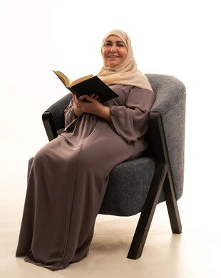 ثقافة التعلم الذاتي، قضاء الوقت بالتعلم والتثقف، الاستمتاع بقراءة الكتب والقصص والروايات، بورترية لإمراة كبيرة بالسن عربية سعودية خليجية محجبة ترتدي العباءة تجلس على كرسي تحمل بيديها كتاب، خلفية بيضاء