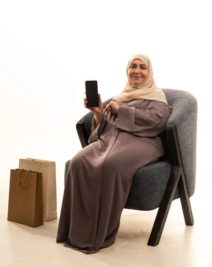 التسوق عبر الانترنت من المحلات والمتاجر الالكترونية، استخدام تطبيقات الهاتف المحمول في شراء المنتجات، بورترية لإمراة كبيرة بالسن عربية سعودية خليجية محجبة ترتدي العباءة تجلس على كرسي بجانبها حقائب التسوق تقوم بعرض الهاتف محمول بشاشه سوداء فارغه، خلفية بيضاء