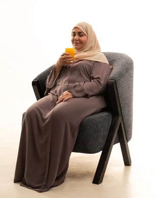 محاولة التخفيف من حرارة الصيف بالمشروب المنعش، الاستمتاع بتناول المشروبات المفيدة اللذيذة، بورترية لإمراة كبيرة بالسن عربية سعودية خليجية محجبة ترتدي العباءة تجلس على كرسي تستمع بشرب العصير الطبيعي البارد، خلفية بيضاء