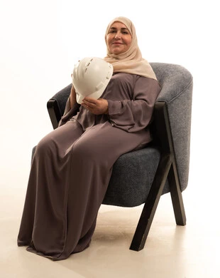 The progress and development of Saudi Arabia at the hands of the sons of the nation, the concept of engineering and construction work, professions and jobs for women, a portrait of an elderly Arab Saudi Gulf woman wearing a hijab and an abaya sitting on a chair holding a helmet in her hands, looking at the camera with gestures of happiness, white background.
