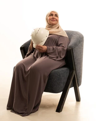 مفهوم الهندسة وأعمال البناء, مهن ووظائف نسائية، تقدم وتطور السعودية على يد أبناء الوطن، بورترية لإمراة كبيرة بالسن عربية سعودية خليجية محجبة ترتدي العباءة تجلس على كرسي تمسك بيديها خوذة, النظر الى شيء ما، خلفية بيضاء