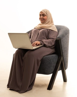 Gestures of integration and focus, merging modern technologies and technology into practical life, accomplishing tasks and work, the concept of remote work, a portrait of an elderly Arab Gulf Saudi woman wearing a hijab and an abaya sitting on a chair using a laptop, white background.