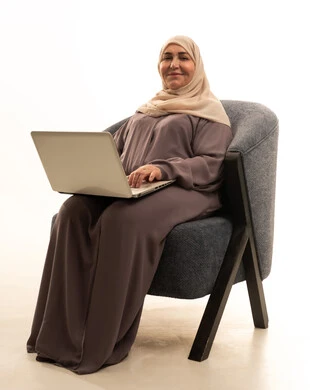 النظر الى الكاميرا بايماءات السعادة و السرور، دمج التقنيات الحديثة والتكنولوجيا في الحياة العملية, انجاز المهام والأعمال, مفهوم العمل عن بعد, بورتريه لإمرأة كبيرة بالسن عربية خليجية سعودية محجبة ترتدي العباءة تجلس على الكرسي تستخدم جهاز الحاسوب المحمول, خلفية بيضاء