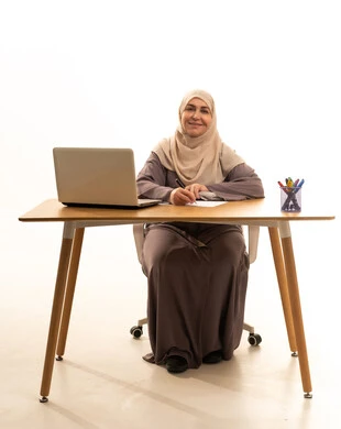 مهنة ووظيفة نسائية، العمل عن بعد، ادارة الأعمال وانجاز المهام المطلوبة، بورتريه لإمراة كبيرة بالسن عربية سعودية خليجية محجبة ترتدي العباءة تجلس على الطاولة تستخدم الحاسوب المحمول و تقوم بكتابة الملاحظات، استخدام جهاز تقني حديث ومتطور، النظر الى الكاميرا بايماءات السعادة و السرور، خلفية بيضاء