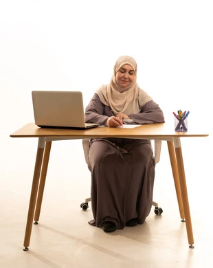 كتابة و تدوين الملاحظات، مهنة ووظيفة نسائية، العمل عن بعد، ادارة الأعمال وانجاز المهام المطلوبة، بورتريه لإمراة كبيرة بالسن عربية سعودية خليجية محجبة ترتدي العباءة تجلس على الطاولة تستخدم الحاسوب المحمول ، استخدام جهاز تقني حديث ومتطور، خلفية بيضاء
