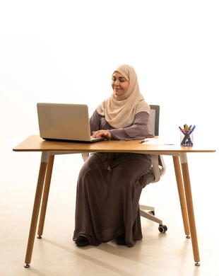 Using a modern and advanced technical device, working remotely, managing business and completing required tasks, a profession and job for women, a portrait of an elderly Arab Saudi Gulf woman wearing a hijab and an abaya sitting at a table using a laptop, white background.