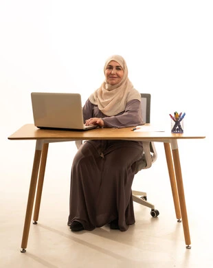 Remote work, business management, and completing required tasks, a profession and job for women, a portrait of an elderly Arab Saudi Gulf woman wearing a hijab and an abaya sitting at a table using a laptop, utilizing a modern and advanced technological device, looking at the camera with gestures of happiness and joy, white background. Remote work, business management, and completing required tasks, a profession and job for women, a portrait of an elderly Arab Saudi Gulf woman wearing a hijab and an abaya sitting at a table using a laptop, utilizing a modern and advanced technological device, looking at the camera with gestures of happiness and joy, white background.