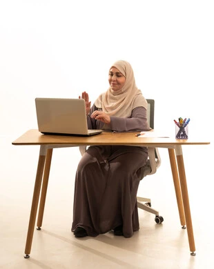 رفع كف الايد بتعابير السلام و التحية، مهنة ووظيفة نسائية، العمل عن بعد، ادارة الأعمال وانجاز المهام المطلوبة، بورتريه لإمراة كبيرة بالسن عربية سعودية خليجية محجبة ترتدي العباءة تجلس على الطاولة تستخدم الحاسوب المحمول ، استخدام جهاز تقني حديث ومتطور، خلفية بيضاء