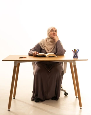Thinking about something, spending time learning and educating oneself, the culture of self-learning, enjoying reading books, stories, and novels, a portrait of an elderly Arab Saudi Gulf woman wearing a hijab and an abaya sitting at a table with a book in front of her, white background.