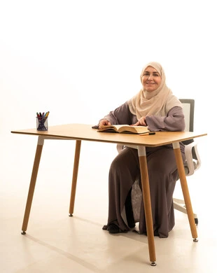 ثقافة التعلم الذاتي، قضاء الوقت بالتعلم والتثقف، الاستمتاع بقراءة الكتب والقصص والروايات،بورتريه لإمراة كبيرة بالسن عربية سعودية خليجية محجبة ترتدي العباءة تجلس على الطاولة امامها كتاب، النظر الى الكاميرا بايماءات السعادة و السرور، خلفية بيضاء