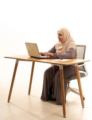 The concept of remote work, integrating modern technologies and technology into professional life, accomplishing tasks and work, a portrait of an elderly Arab Gulf Saudi woman wearing an abaya and hijab sitting at a table using a laptop, white background.