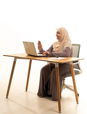 استخدام جهاز الحاسوب المحمول، دمج التقنيات الحديثة والتكنولوجيا في الحياة العملية, انجاز المهام والأعمال, مفهوم العمل عن بعد, بورترية لامراة عربية خليجية سعودية كبيرة في السن ترتدي العباءة و الحجاب تجلس على الطاولة ترفع كف يدها بايماءات القاء التحية والسلام, خلفية بيضاء