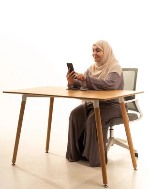 Using a modern and advanced technical device, browsing social media sites, chatting with friends and relatives via mobile, a portrait of an elderly Arab Saudi Gulf woman wearing a hijab and an abaya sitting at a table holding a mobile phone in her hand, white background.
