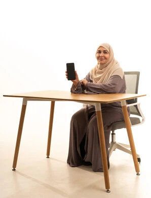 استخدام الأجهزة التقنية الحديثة للتسلية والترفيه, النظر إلى الكاميرا بإيماءات السعادة و السرور, بورتريه لإمراة كبيرة بالسن عربية سعودية خليجية محجبة ترتدي العباءة تجلس على الطاولة تمسك بيدها الهاتف المحمول, عرض شاشة سوداء فارغة عبر الجوال, خلفية بيضاء