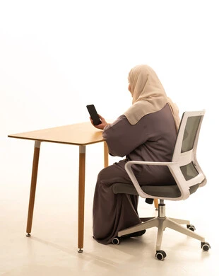 تصفح مواقع التواصل الاجتماعي، الدردشة مع الأصدقاء والأقارب عبر الجوال، استخدام جهاز تقني حديث ومتطور،بورتريه من الخلف لإمراة كبيرة بالسن عربية سعودية خليجية محجبة ترتدي العباءة تجلس على الطاولة تمسك بيدها الهاتف المحمول، خلفية بيضاء