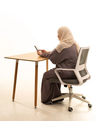 Chatting with friends and relatives via mobile, using a modern and advanced technical device, browsing social media sites, a portrait from behind of an elderly Arab Saudi Gulf woman wearing a hijab and an abaya sitting at a table holding a mobile phone in her hand, white background.