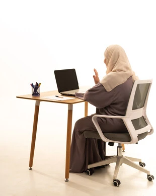 Integrating modern technologies and technology into practical life, accomplishing tasks and work, the concept of remote work, and a side portrait of an elderly Saudi Gulf Arab woman wearing an abaya and hijab sitting at a table raising her hand in gestures of greeting and peace, using a laptop, white background.