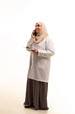مفهوم الطب والرعاية الصحية، دمج التقنيات الحديثة في الحياة العملية، مهن ووظائف سعودية,بورتريه لطبيبة عربية خليجية سعودية كبيرة في السن ترتدي المعطف الطبي تحمل دفتر سجلات المرضى تقوم باجراء مكالمة هاتفية عبر الهاتف المحمول, الجسم بالطول الكامل، خلفية بيضاء